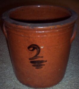 Whitewater two-gallon crock with ear handles and a painted "2" with three underlines. The color is deep red/orange. While most Whitewater clay is light cream colored there are some outliers. The dark color is probably caused by iron content exposed to air. It could from clay mined near the surface or clay that was exposed to air for a time before being turned.